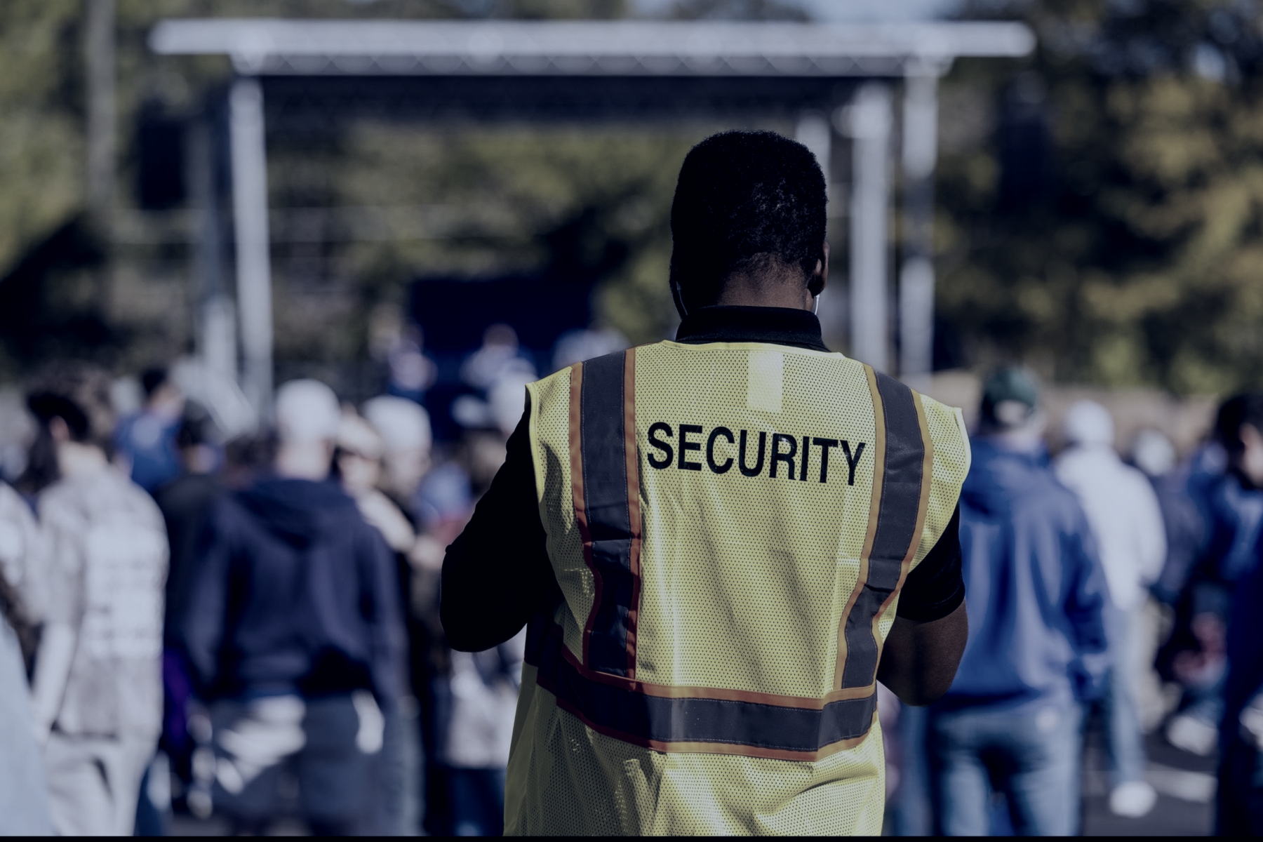 An event safety officer in a hi-vis vest lettered SECURITY, seen from behind, watching an outdoor crowd in front of a stage gantry.