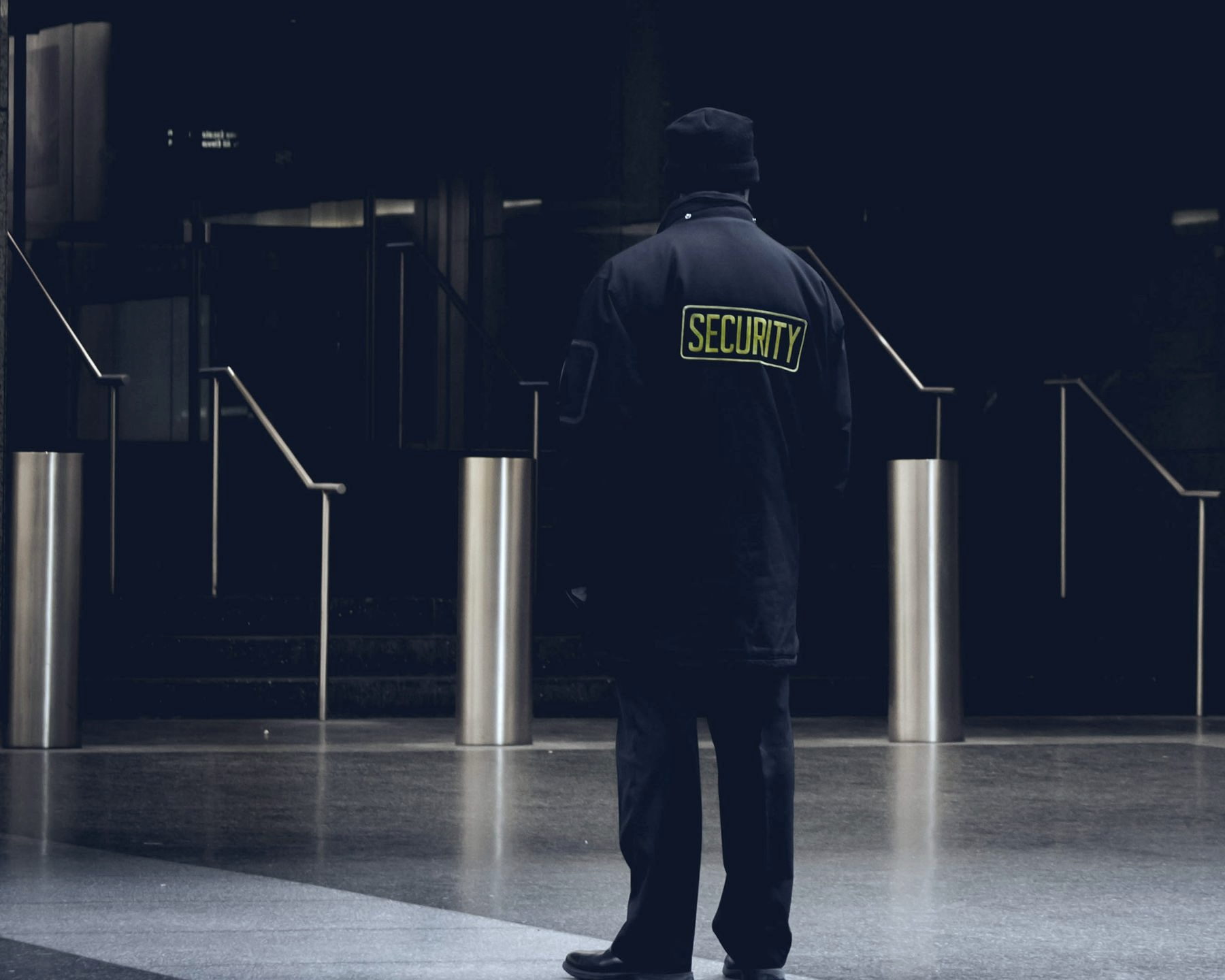 A security officer seen from behind, SECURITY lettered in yellow across the back of his dark coat, standing among brushed steel bollards on the forecourt of an office building entrance at night.