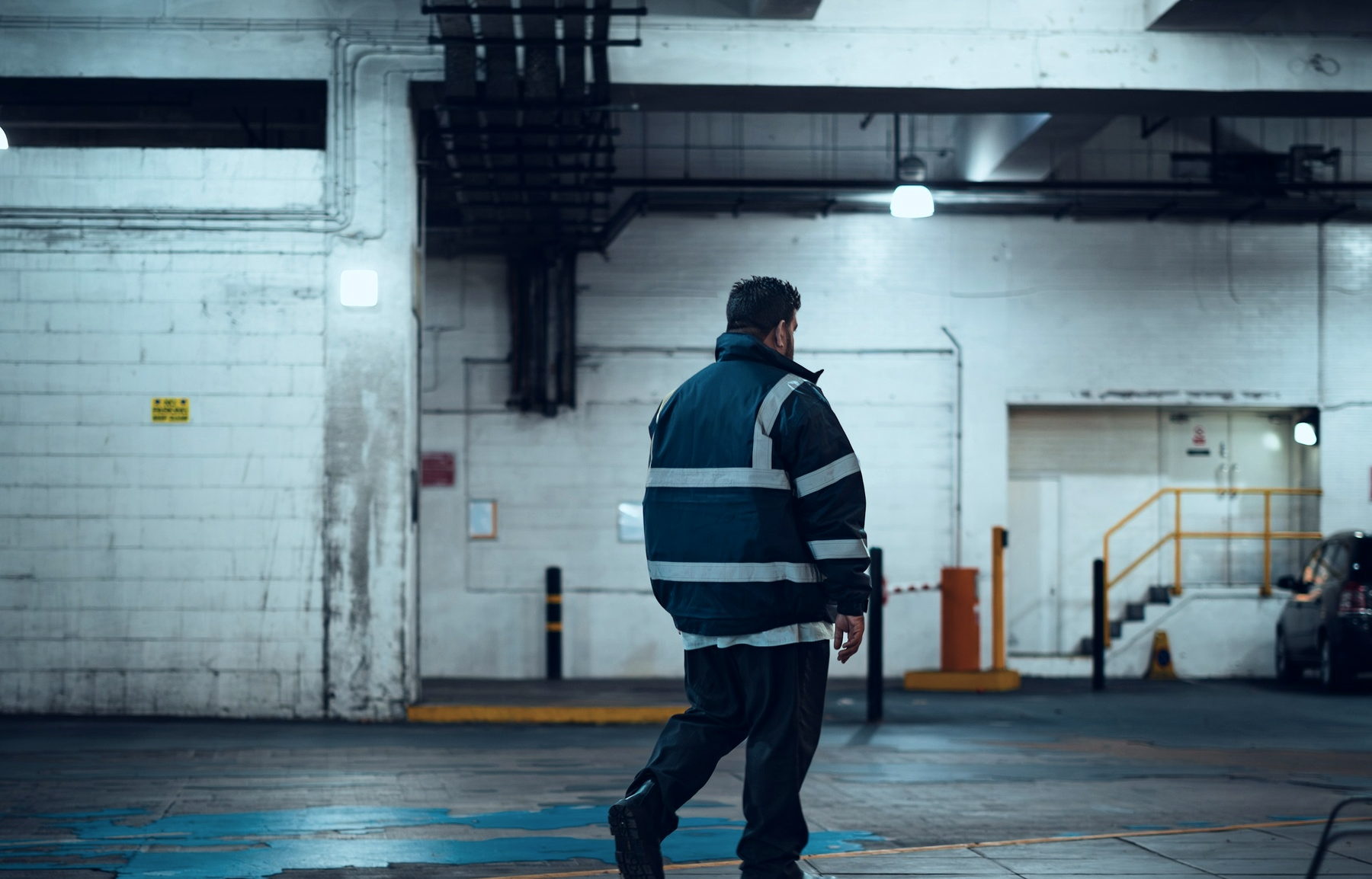 A patrol officer in a navy jacket with reflective bands, seen from behind, walking through an underground car park at night, with a parked car at the far right.
